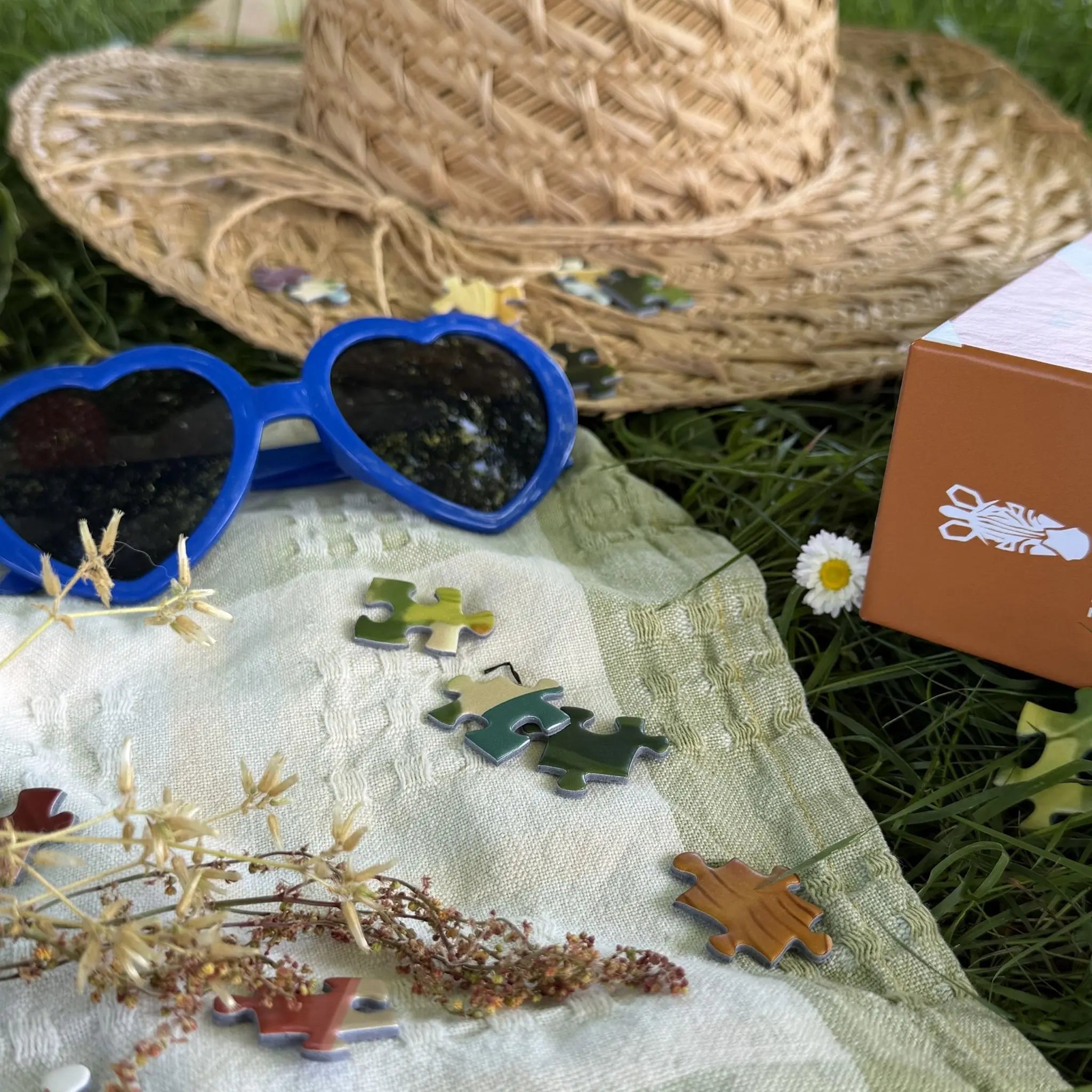 Ambiance estivale autour du puzzle adulte ZEBRESQUE avec pièces de puzzle, lunettes cœur bleues, chapeau de paille et tissu de pique-nique dans l’herbe.