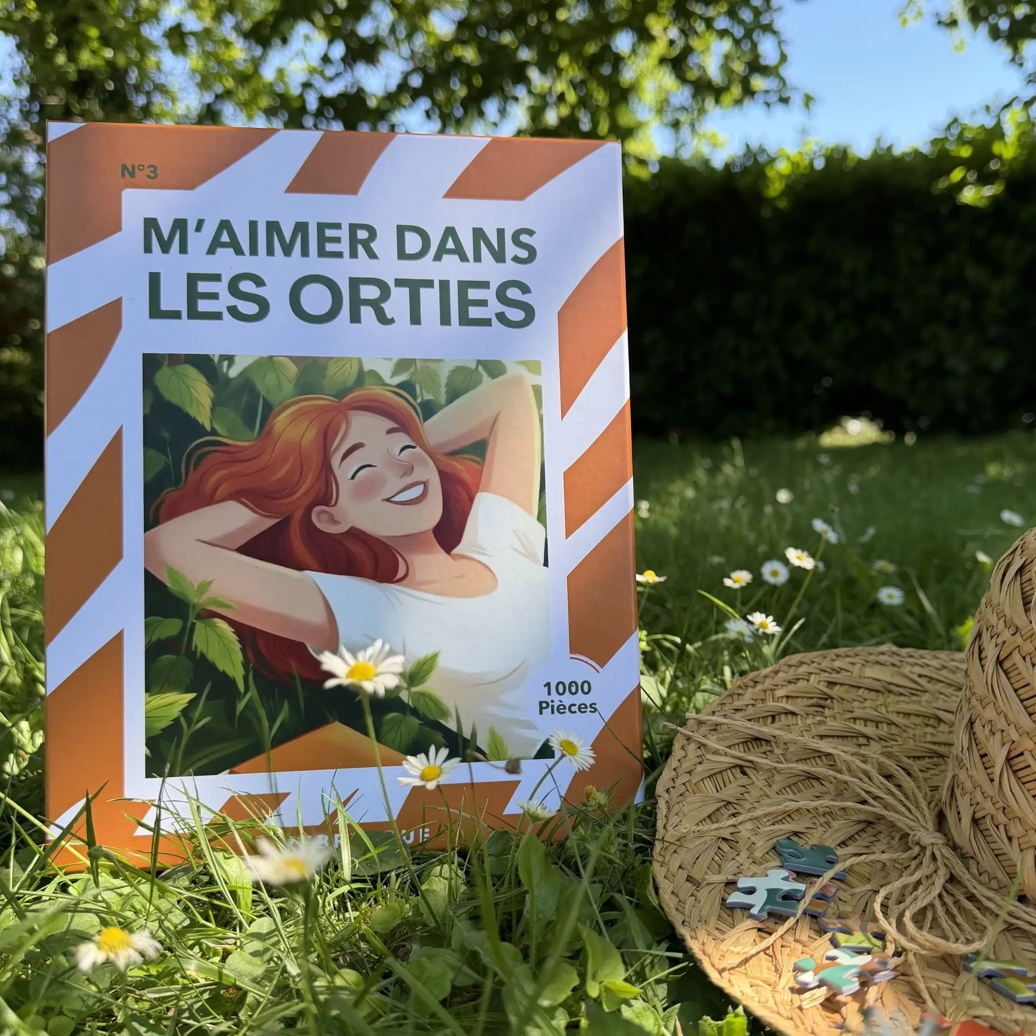 Boîte du puzzle adulte 1000 pièces ZEBRESQUE “M’aimer dans les orties” photographiée dans un jardin ensoleillé avec des pâquerettes, de l’herbe et un panier en osier. Illustration féminine et nature dans une ambiance calme, contemplative et déconnexion.