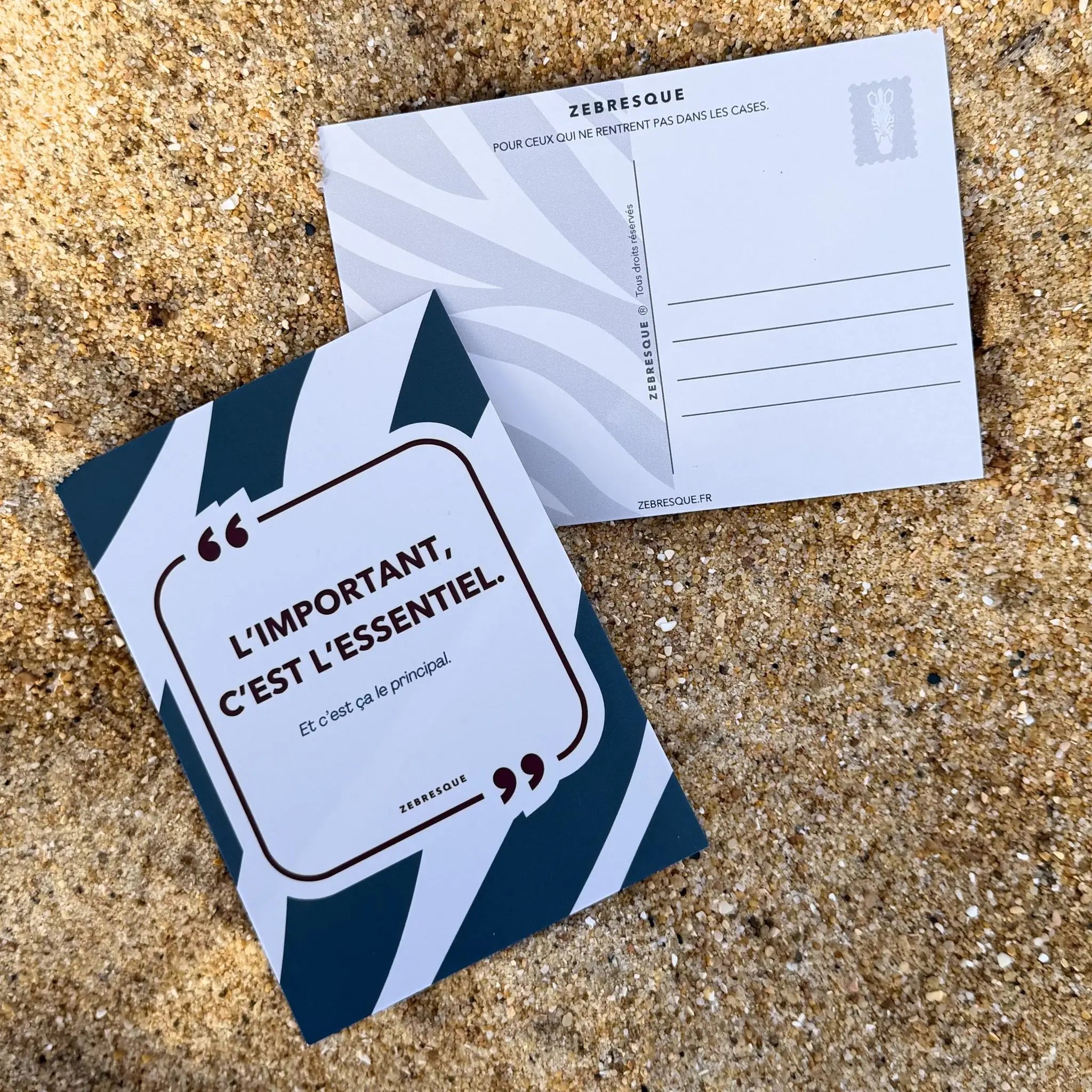 Carte postale minimaliste ZEBRESQUE “L’important, c’est l’essentiel.” posée sur le sable. Objet graphique inspiré des pensées lentes, des pauses volontaires et des esprits qui cherchent du calme au milieu du bruit.