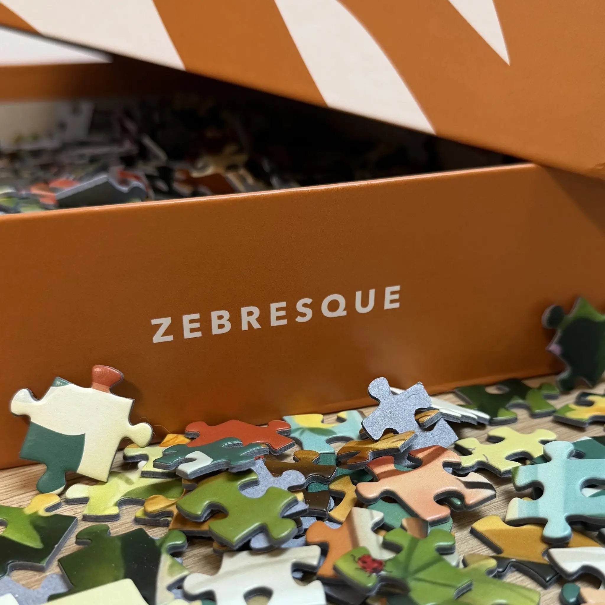 Détail de la boîte du puzzle adulte 1000 pièces ZEBRESQUE ouverte avec des pièces illustrées aux tons verts et naturels éparpillées sur une table en bois dans une ambiance calme et anti-stress.
