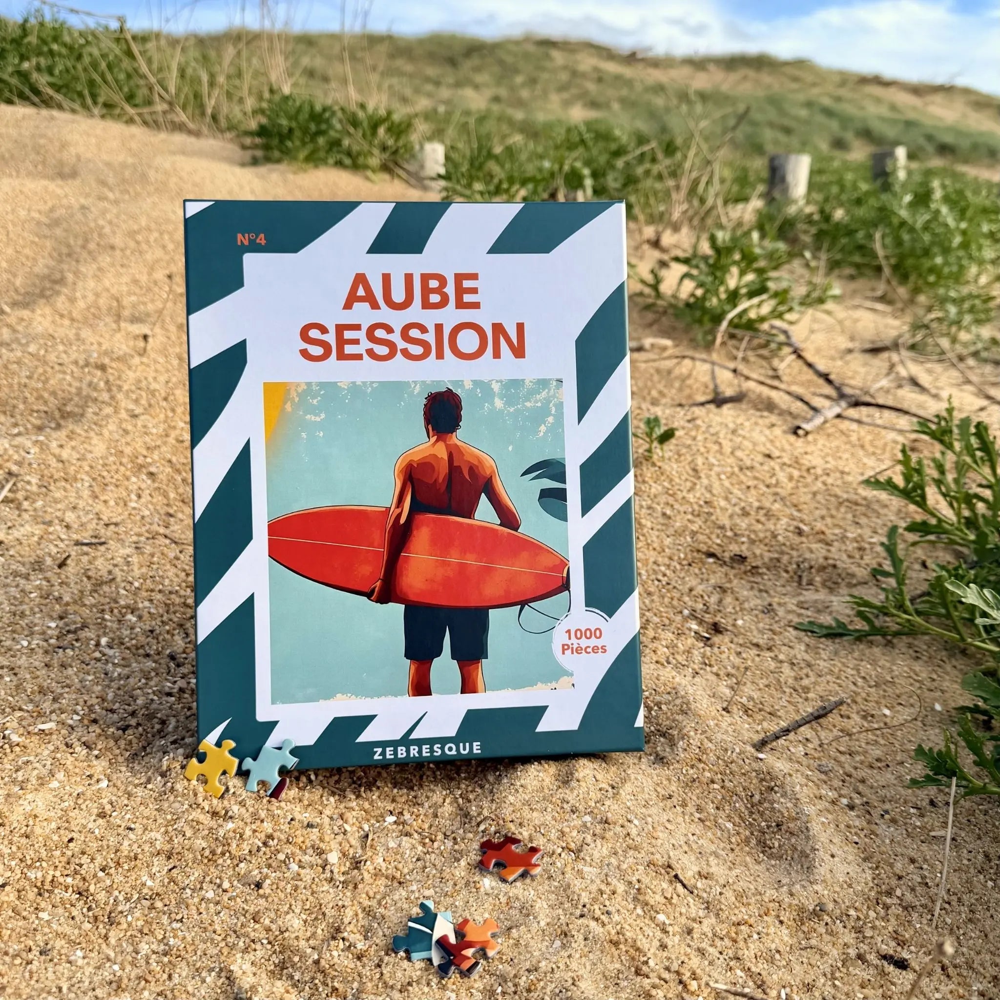 Puzzle adulte 1000 pièces ZEBRESQUE « Aube Session » posé sur le sable face à l’océan dans les Landes, avec illustration d’un surfeur au lever du jour dans une ambiance slow life inspirée des sessions de surf matinales