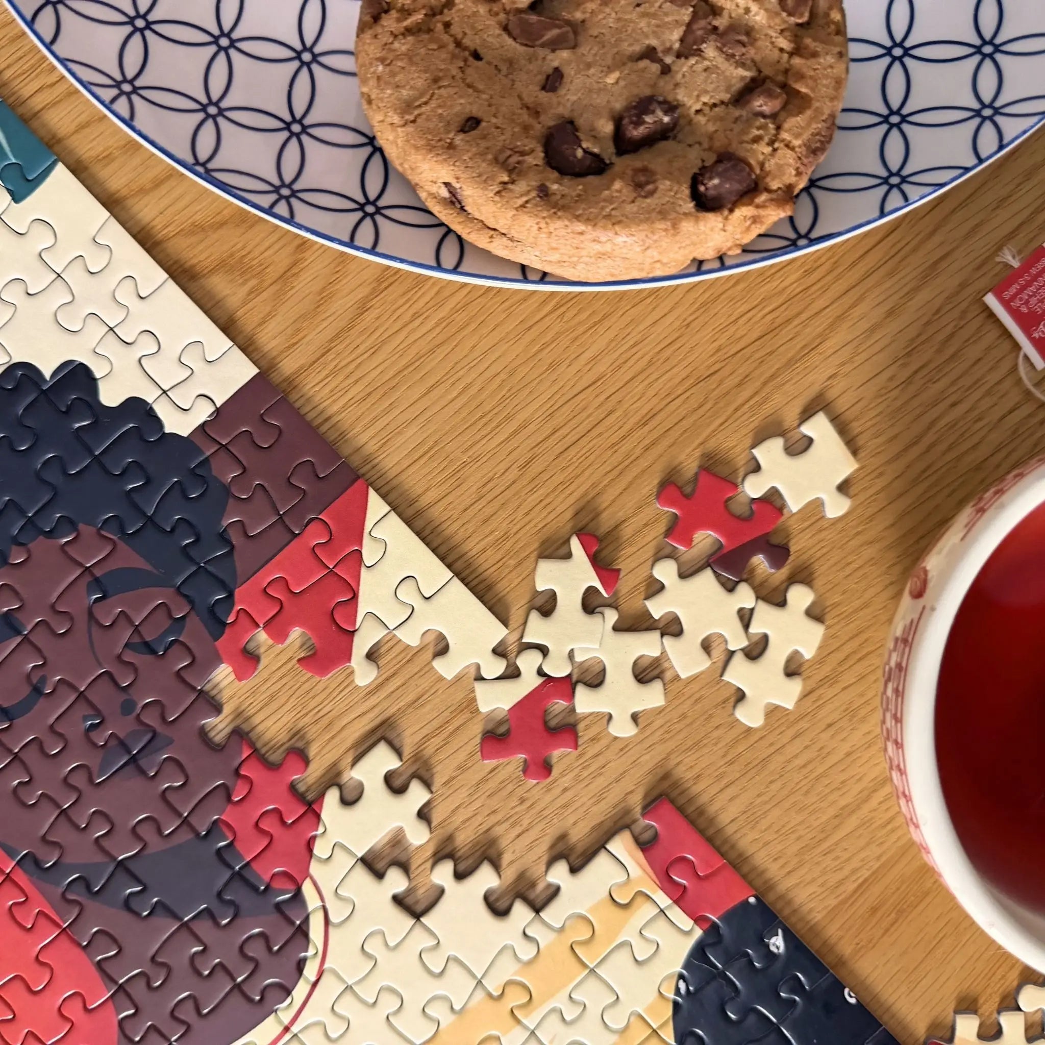 Détail d’un puzzle DECLARER SA FLEMME pour adulte avec une femme endormie de 1000 pièces ZEBRESQUE avec pièces dispersées sur une table en bois, tasse de thé rouge et cookie dans une ambiance cosy slow life.