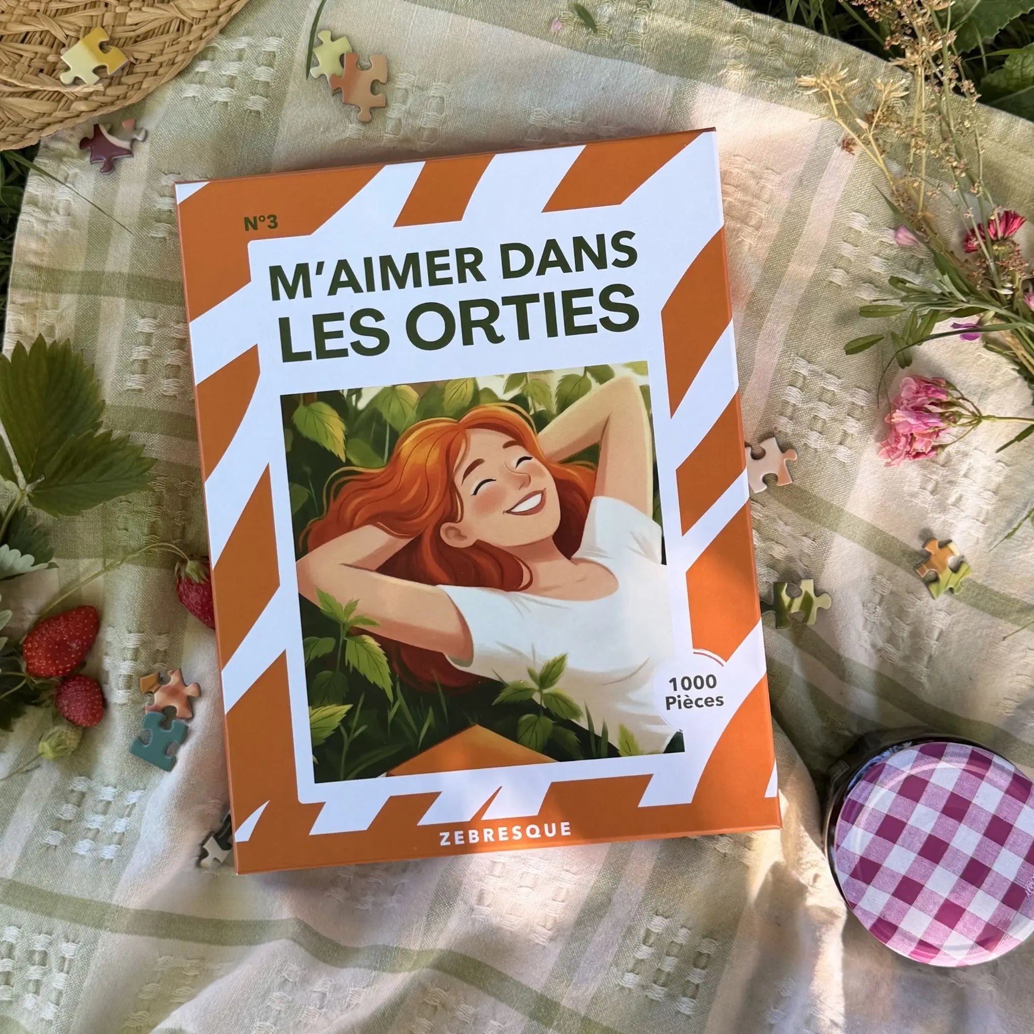 Boîte du puzzle adulte 1000 pièces ZEBRESQUE “M’aimer dans les orties” posée sur une couverture de pique-nique avec fleurs, fraises et pièces de puzzle dans une ambiance estivale, calme et slow life