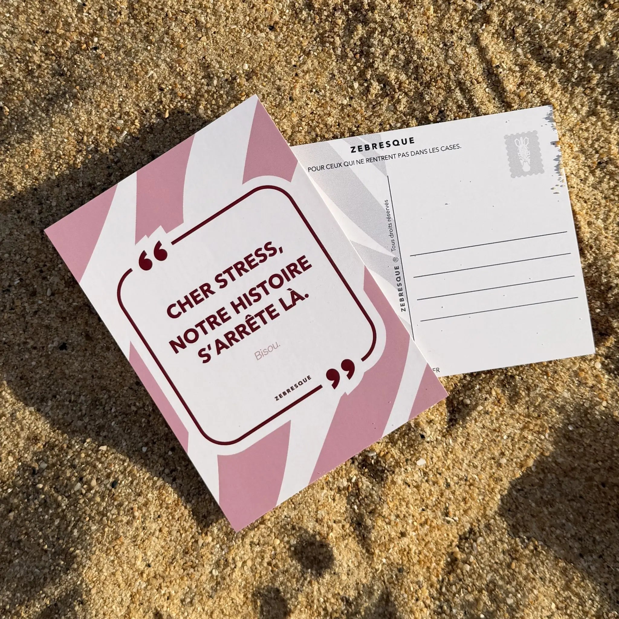 Carte postale minimaliste ZEBRESQUE “L’important, c’est l’essentiel.” posée sur le sable. Objet graphique inspiré des pensées lentes, des pauses volontaires et des esprits qui cherchent du calme au milieu du bruit.