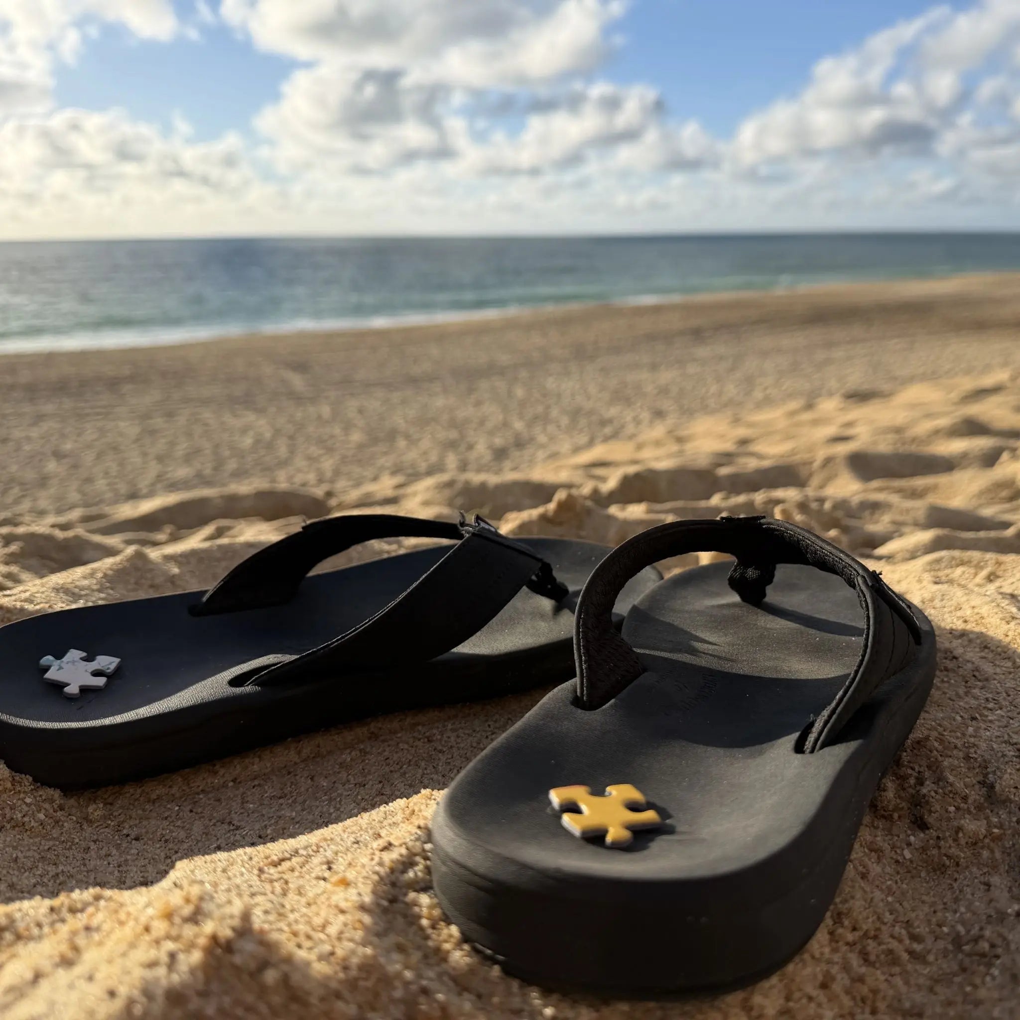 Tongs noires posées dans le sable face à l’océan avec pièces de puzzle ZEBRESQUE dans une ambiance slow life inspirée du surf, des plages landaises et des sessions matinales contemplatives
