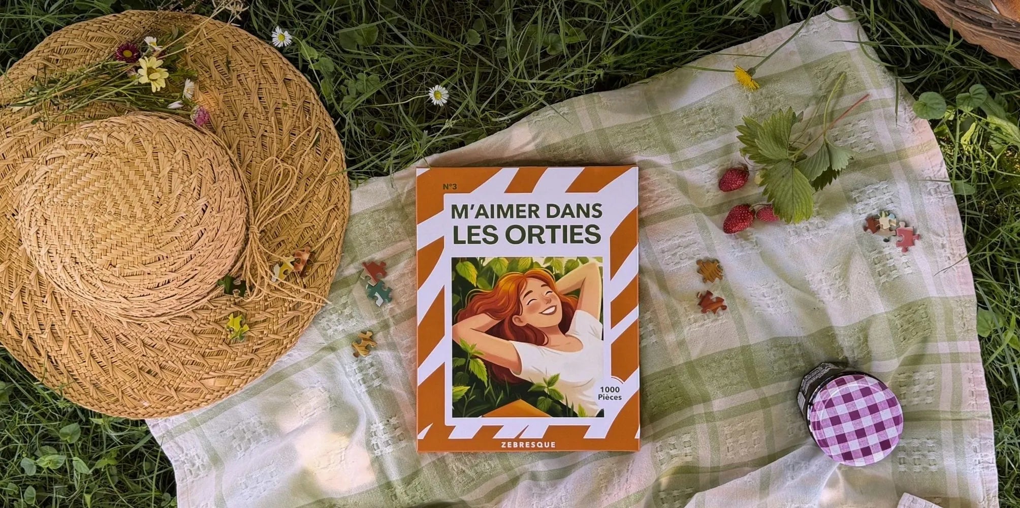 Puzzle adulte 1000 pièces ZEBRESQUE “M’aimer dans les orties” fabriqué en France posé sur un plaid dans l’herbe avec chapeau de paille, fraises sauvages et ambiance pique-nique estivale apaisante.